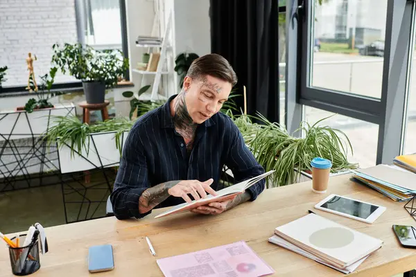 Genç bir adam yeşilliklerle çevrili bir masada otururken düşünceli bir şekilde bir kitapla meşgul olur..