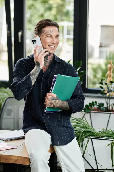 stock image A young professional chats on the phone while holding colorful notebooks in a bright workspace.