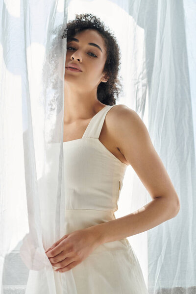 A fashionable woman stands thoughtfully beside sheer curtains, illuminated by warm sunlight.