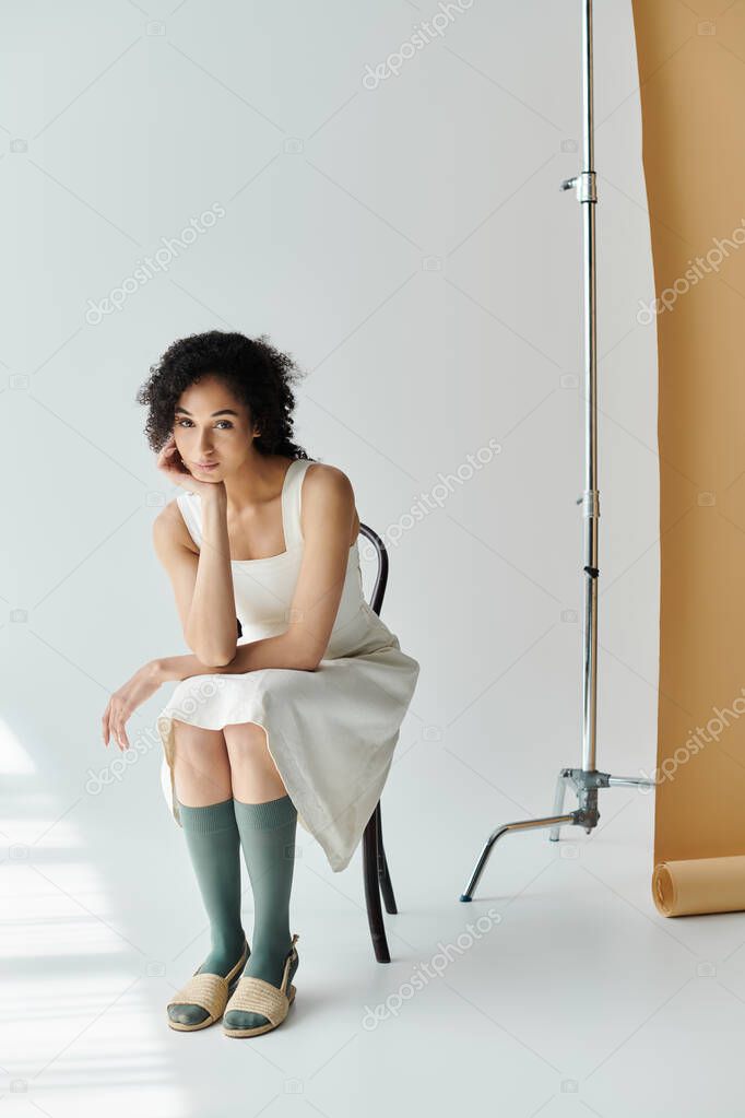 A fashionable woman sits thoughtfully on a chair, showcasing her chic outfit in soft light.