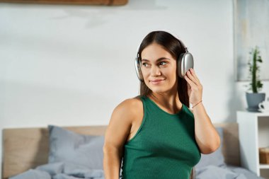 Kısa boylu esmer bir kadın rahat yatak odasında müzik dinliyor, evdeki huzurlu zamanlarından zevk alıyor..