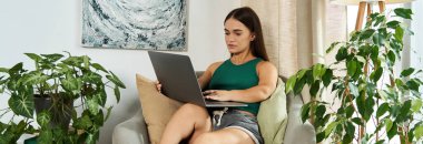 Kısa boylu esmer bir kadın rahat bir şekilde oturma odasında oturuyor, laptopuna odaklanmış, çevresi yemyeşil..