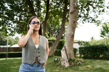 Şık bir yeleği ve güneş gözlüklü genç bir kadın telefonunda sohbet ediyor, güneşli bir bahçenin güzelliğine dalmış..