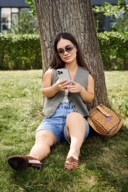 A young woman with brunette hair relaxes in the sun, sitting under a tree while focused on her smartphone, embracing the moment. clipart