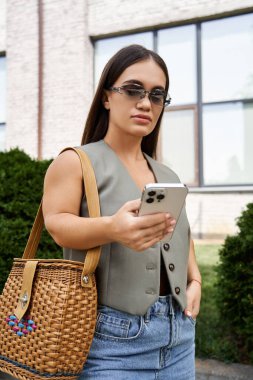 Kısa boylu esmer bir kadın dışarıda günlük kıyafetleriyle dikiliyor. Etrafında güneş parlarken telefonuna odaklanıyor..