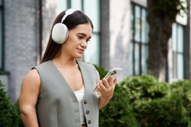 Açık havada akıllı telefonuna bakarken kulaklıklı genç bir esmer kadın gülümsüyor..