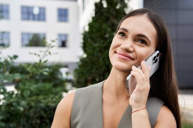 A cheerful young woman with short stature engages in a lively phone conversation, surrounded by urban greenery and bright sunshine. clipart