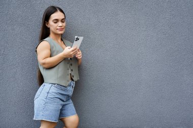 Kısa boylu, esmer bir kadın telefonunu kontrol ederken dışarıda vakit geçirmekten zevk alıyor. Rahat bir hava yayıyor..