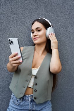 A young brunette woman with a short stature relaxes outdoors, listening to music on her phone, smiling as she engages with the moment. clipart