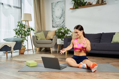 Evde kısa boylu esmer bir kadın, yoga minderinde laptopunun talimatlarını hararetle uyguluyor..
