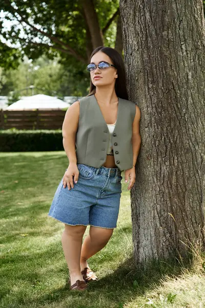 stock image A petite brunette smiles while leaning against a sturdy tree, soaking in the warmth of a sunny day outdoors.