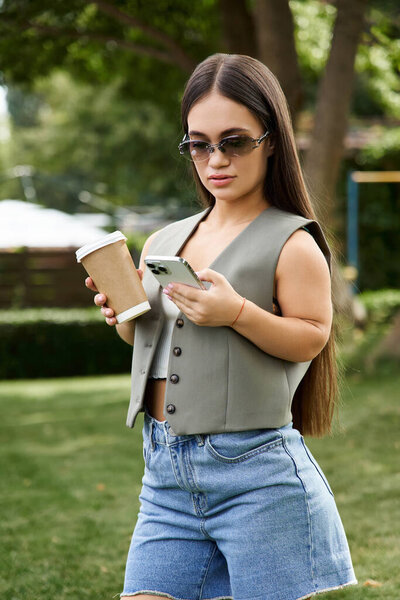 A young brunette woman with short stature stands in a sunlit outdoor space, sipping coffee and texting on her phone, relishing her time.