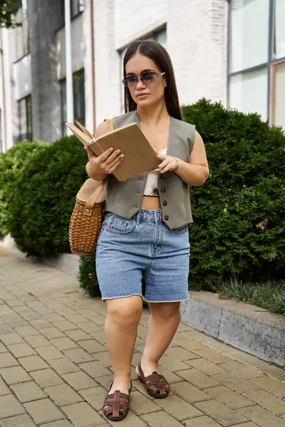 stock image The young brunette woman with short stature enjoys a sunny outdoor moment, engrossed in her book while strolling along a picturesque path.