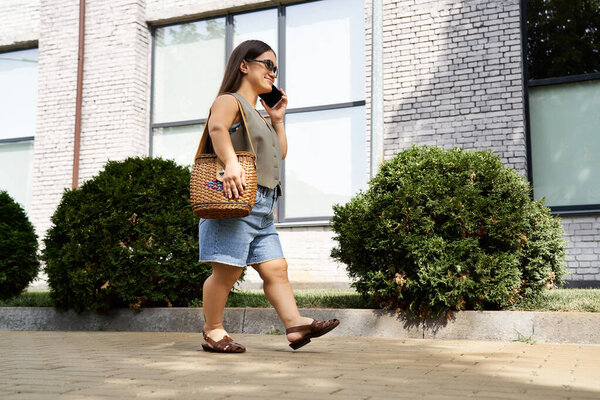 A petite brunette woman strolls along a tree-lined path, engaged in a lively phone conversation, radiating joy.