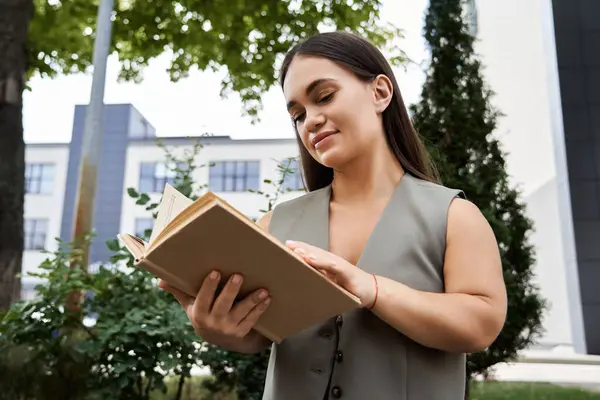 Stock image A young brunette woman with a joyful expression enjoys reading a book in an inviting outdoor space surrounded by greenery.