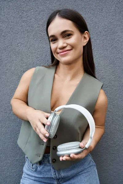 stock image A young brunette woman stands outdoors, cheerfully holding her headphones, radiating joy and style under the sun.