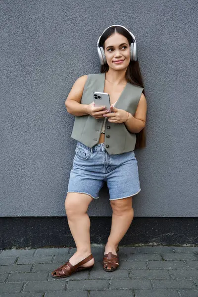 stock image A young brunette woman with a cheerful demeanor relaxes outdoors, wearing stylish summer clothes and headphones, enjoying her music.