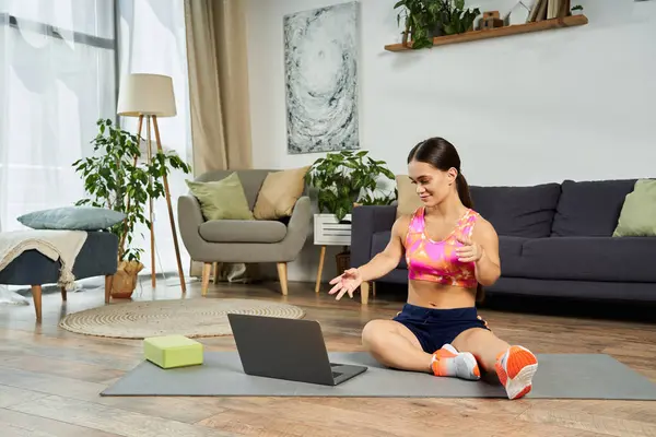 stock image A young brunette woman with a short stature exercises at home, enthusiastically following instructions from her laptop on a yoga mat.