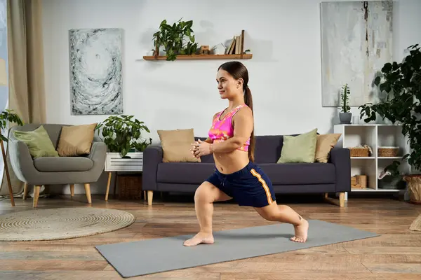 stock image A young brunette woman with a short stature exercises at home, practicing lunges on a mat in her cozy living room filled with plants.