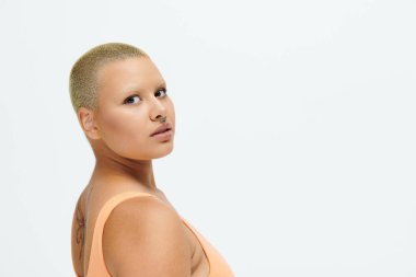 Genç kel bir kadın, basit bir mekanda kendine güven ve tarz yayarak renkli giysiler sergiliyor..