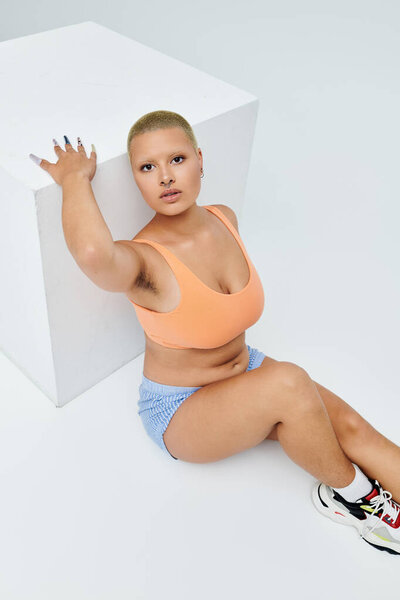 A young bald woman in vibrant attire strikes a bold pose beside a geometric white structure.