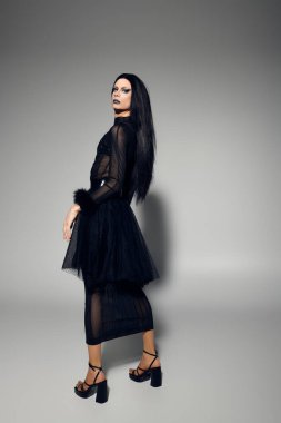 Şık bir travesti, gri bir arkaplana karşı özgüven ve sanatsal bir ortam sergiliyor..
