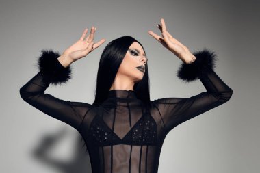 Büyüleyici bir travesti zarif bir şekilde siyah bir elbise içinde gri bir arkaplana karşı poz verir..