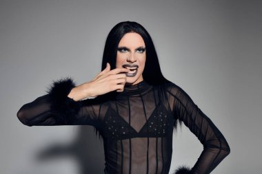 Gösterişli, siyah bir elbise içinde zarif bir travesti, soluk benizli bir griye karşı güven ve sanatsal bir tavır sergiliyor..