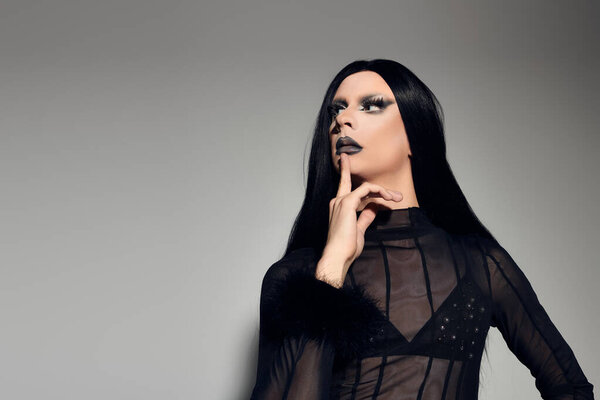 A fashionable drag queen exudes confidence in a stylish black outfit, striking a pose in a studio setting.