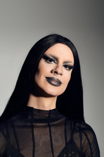 A confident drag queen in a striking black outfit poses elegantly against a smooth grey backdrop.