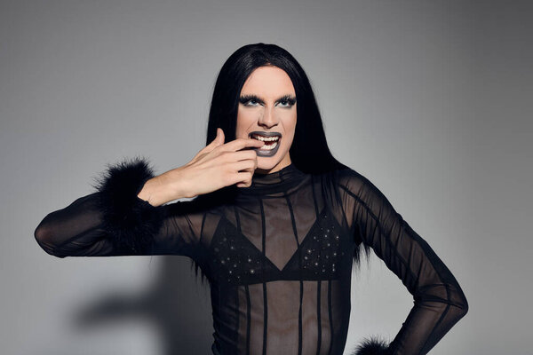 In a sleek black outfit, an elegant drag queen showcases confidence and artistry against a muted grey.