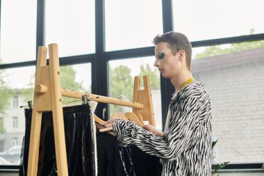 Cinsiyet değişkenliği olan bir moda tasarımcısı, parlak bir stüdyo ortamında cüretkar tarzlarını sergileyerek giysi seçer..