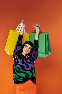 A young woman showcases her colorful style with a bright sweater and shopping bags against a vivid orange background. clipart