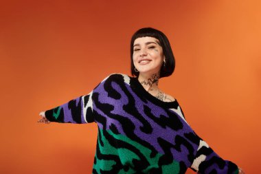 A cheerful woman with a stylish haircut and vibrant tattoos strikes a playful pose in a colorful sweater. clipart