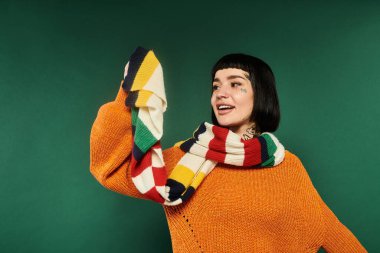 Kısa saçlı ve dövmeli genç bir kadın, yeşil bir kazak giyerken sevinçten havalara uçuyor..