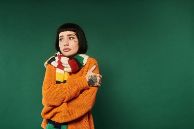 A young woman with tattoos and short hair stands warmly wrapped in a colorful sweater against a green backdrop. clipart