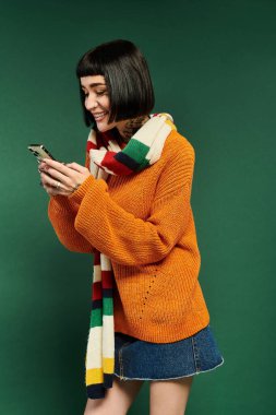 A young woman with short hair and tattoos smiles while checking her phone, dressed warmly in a cozy sweater. clipart