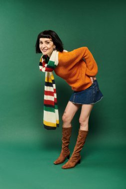 A cheerful young woman with short hair and tattoos poses playfully in a cozy sweater against a green backdrop. clipart