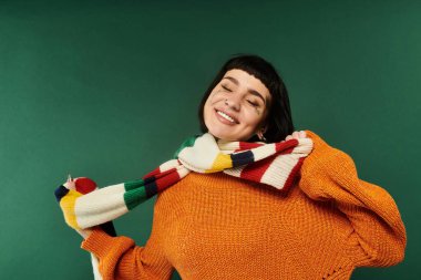 Kısa saçlı ve dövmeli genç bir kadın canlı bir süveter ve eşarp giyerken sıcak bir şekilde gülümsüyor..