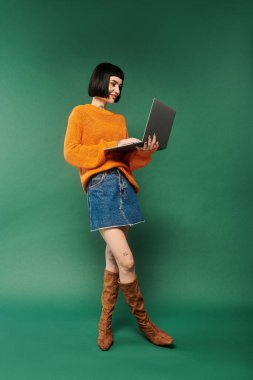 A young woman with short hair and tattoos poses confidently in a cozy sweater and using laptop against a green backdrop. clipart