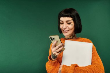 Canlı dövmeleri ve kısa saçlı genç bir kadın telefonunu kontrol ederken gülümsüyor, rahat bir kazak giyiyor..