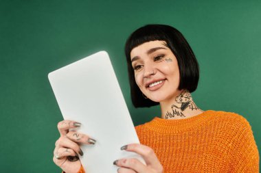 Rahat bir süveter ve dövmeler içinde gülümseyen genç bir kadın yeşil bir zemine sıcaklık saçıyor..