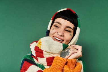 Kısa saçlı ve dövmeli neşeli genç bir kadın rahat bir süveter ve atkıyla poz veriyor, sıcaklık ve neşe saçıyor..