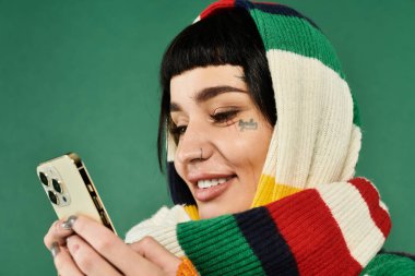 Kısa saçlı ve dövmeli genç bir kadın yeşil arka planda rahat bir kazak ve eşarpla telefonunu kontrol ederken gülümsüyor..
