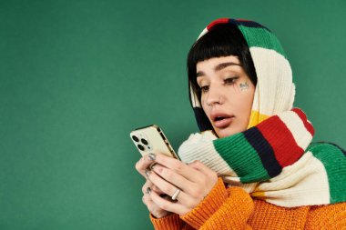 Renkli bir kazak ve atkı giymiş genç bir kadın telefonuna özenle bakıyor, sıcaklığını ve tarzını yansıtıyor..