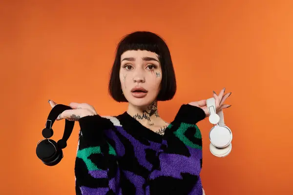 stock image A young woman with short hair and tattoos poses confidently in a bright sweater and holding headphones on a vibrant orange background.