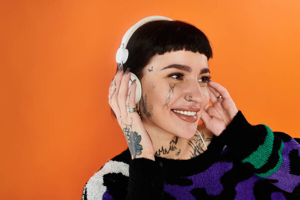 Молодая женщина в ярком свитере и наушниках улыбается, позируя на оранжевом фоне.