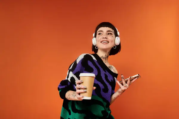 stock image A young woman with short hair and tattoos smiles while holding a drink and phone, immersed in music.