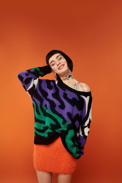 Молодая женщина с короткими волосами и татуировками излучает радость в красочном свитере на ярком оранжевом фоне.