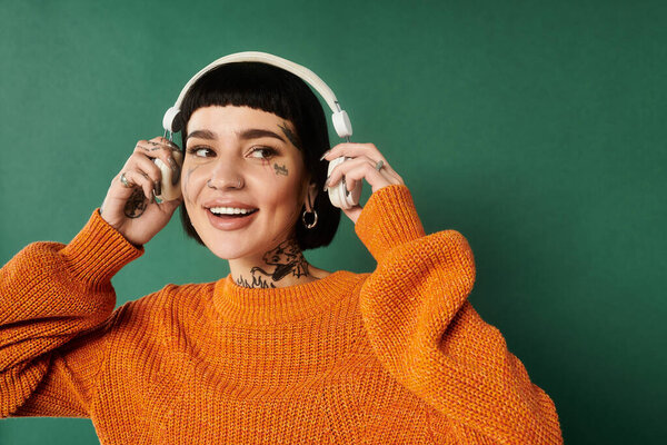 A cheerful young woman with short hair and tattoos smiles, wearing cozy attire while listening to music.